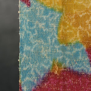 May include: Close-up of a colourful woven textile with a vibrant pattern. The fabric features a mix of blue, yellow, and red hues, with a textured weave. The edges are unfinished, revealing the raw texture of the material.