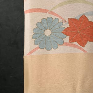 May include: Close-up of a textile with a floral pattern. The design features a light blue flower and a red maple leaf with gold outlines, set against a cream and peach background. The fabric has a textured appearance.