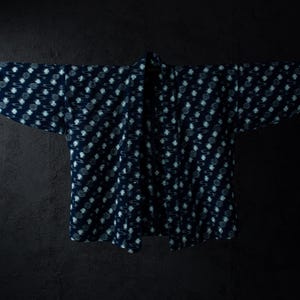 May include: A dark blue indigo-dyed jacket featuring a repeating white floral and circular pattern across the fabric. The garment has wide sleeves, an open front design, and a straight hemline, displayed hanging horizontally on a thin rod.