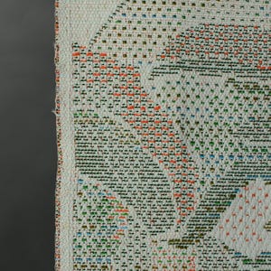 May include: Close-up of a woven textile with a geometric pattern. The design features a repeating pattern of white, orange, green, and blue squares and lines. The edges are finished with a white border.