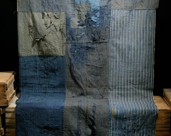 May include: A large patchwork quilt made of various shades of blue and gray fabric. The quilt is made of different sized squares and rectangles, and some of the fabric has been worn and faded. The quilt is folded in half and is sitting on a wooden surface.