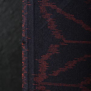 Puede incluir: Primer plano de una tela azul oscuro con un patrón tejido en tonos rojos. El textil tiene una apariencia texturizada, con el diseño rojo creando líneas diagonales y en zigzag en la superficie. La tela se utiliza probablemente para ropa o decoración del hogar.