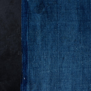May include: A rectangular piece of fabric dyed in a deep indigo blue color. The textile features a visible woven texture with a slightly uneven, organic appearance throughout the surface, showcasing a rich, saturated pigment and a vertical grain.
