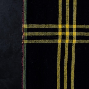 Peut inclure: Un tissu noir présentant un motif à carreaux jaune avec des lignes horizontales et verticales qui se croisent. Le bord gauche du textile affiche une fine bordure multicolore avec des fils verts, rouges et jaunes visibles sur la lisière.