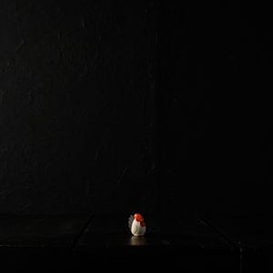 Puede incluir: Pequeña figura de gallina de cerámica blanca con cresta y barbilla rojas. La figura está centrada sobre una superficie de madera oscura, sobre un fondo negro. La iluminación resalta los detalles.
