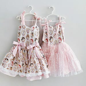 May include: Two dresses hanging on white hangers. One dress is white with a pattern of princess portraits, pink bows, and a lace trim. The other is a pink tulle dress with white polka dots.