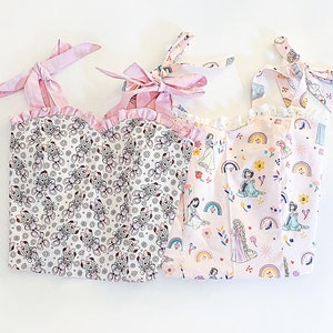 May include: Three pink and white sleeveless tops with different patterns. The first top has a small floral pattern with a brown and white design. The second top has a pink and white pattern with a rainbow and princesses. The third top has a pink and white pattern with a rainbow and princesses.