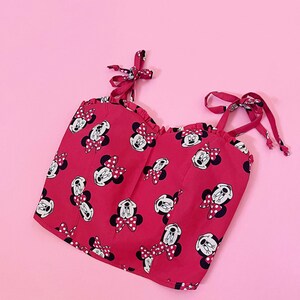 May include: A red crop top with a Minnie Mouse print and white polka dots. The top has thin straps that tie at the shoulders.