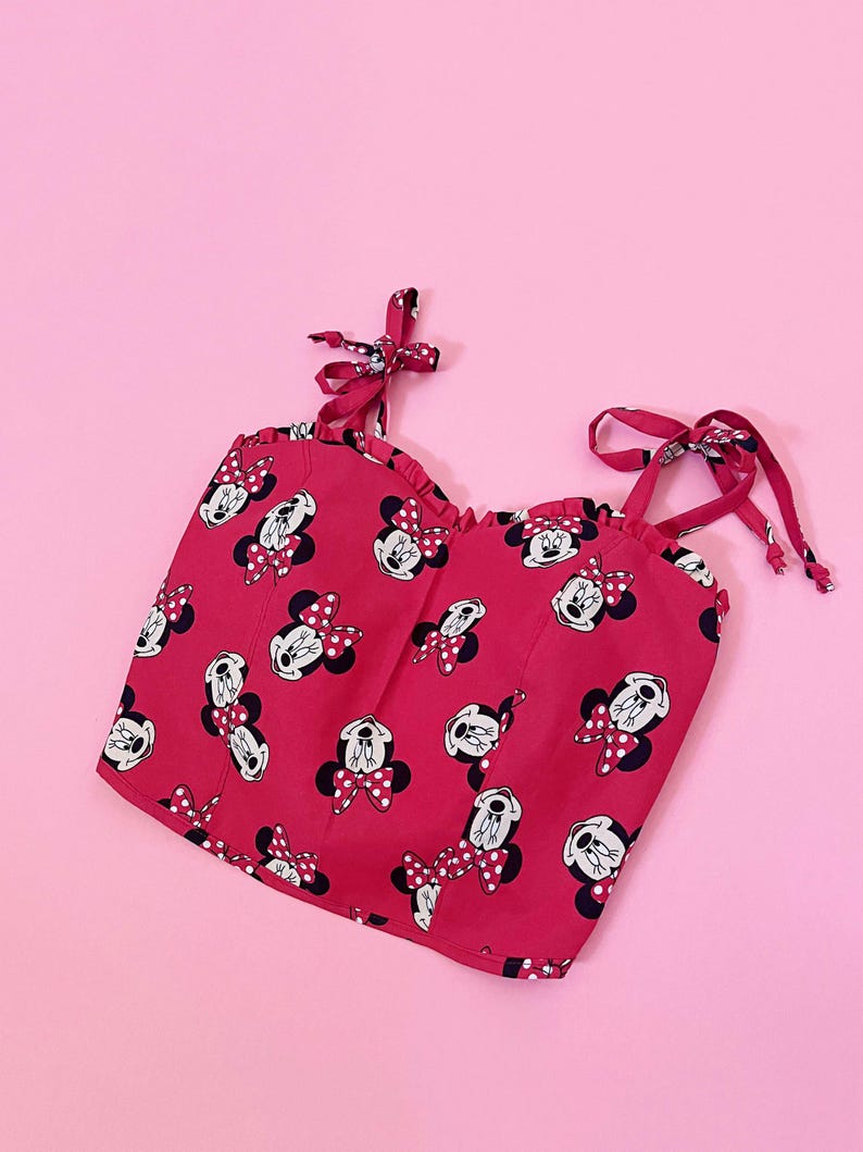 May include: A red crop top with a Minnie Mouse print and white polka dots. The top has thin straps that tie at the shoulders.