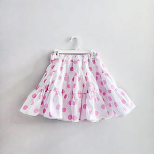 May include: A white skirt with a pink polka dot pattern, hanging on a white hanger. The skirt has a tiered, ruffled design. The skirt is made of a lightweight fabric, suitable for warm weather.