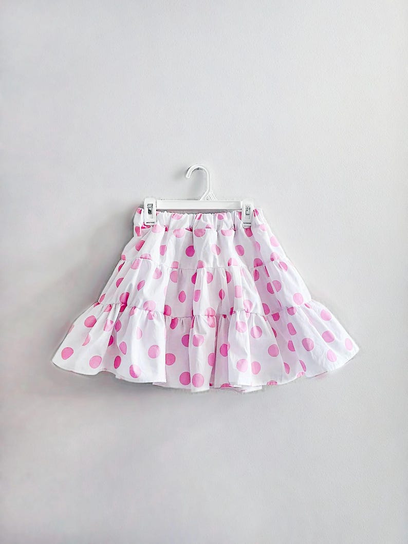 May include: A white skirt with a pink polka dot pattern, hanging on a white hanger. The skirt has a tiered, ruffled design. The skirt is made of a lightweight fabric, suitable for warm weather.