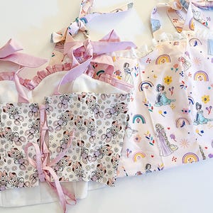 May include: Three pink fabric tops with princess character prints. The top on the left has a white background with a floral pattern and pink straps. The middle top has a white background with a floral pattern and pink straps. The top on the right has a pink background with a rainbow and princess character print.
