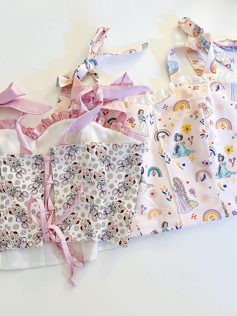 May include: Three pink fabric tops with princess character prints. The top on the left has a white background with a floral pattern and pink straps. The middle top has a white background with a floral pattern and pink straps. The top on the right has a pink background with a rainbow and princess character print.