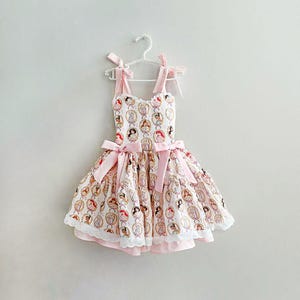 May include: A light pink and white dress with a princess pattern, hanging on a white hanger. The dress features pink bows on the straps and waist, and a white lace trim at the hem. The dress is designed for a child.