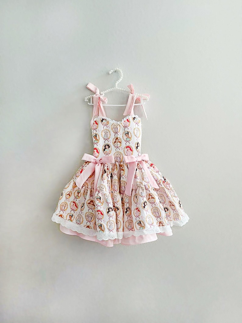 May include: A light pink and white dress with a princess pattern, hanging on a white hanger. The dress features pink bows on the straps and waist, and a white lace trim at the hem. The dress is designed for a child.