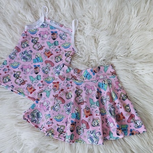 May include: A pink tank top and skater skirt set featuring a colorful cartoon character print. The tank top has white straps and trim. The skirt has a flared design. The set is displayed on a white fluffy surface.