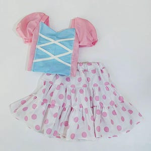 May include: A two-piece outfit comprising a pink and blue top and a white skirt with pink polka dots. The top has puffed sleeves and a blue bodice with white criss-cross detailing. The skirt is tiered, offering a playful, feminine aesthetic.