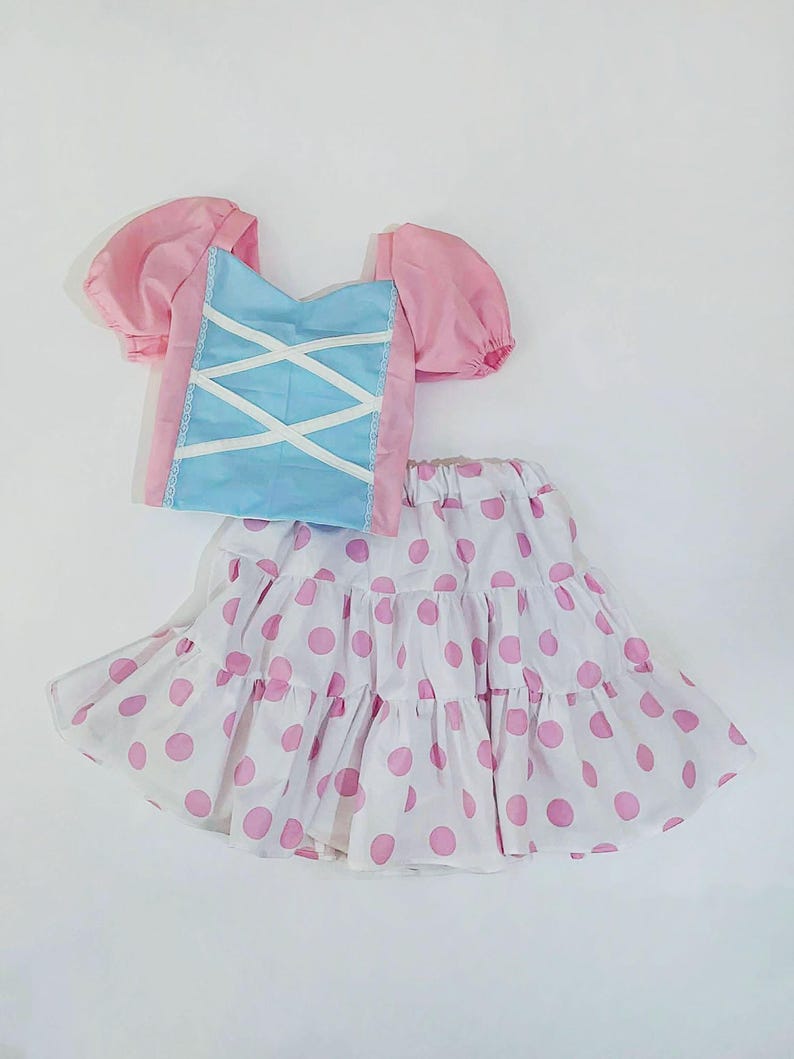 May include: A two-piece outfit comprising a pink and blue top and a white skirt with pink polka dots. The top has puffed sleeves and a blue bodice with white criss-cross detailing. The skirt is tiered, offering a playful, feminine aesthetic.