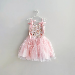 May include: A pink dress with a princess-themed bodice and a layered tulle skirt. The bodice features a repeating pattern of princess portraits. The dress has pink ribbon straps and is hanging on a white hanger.