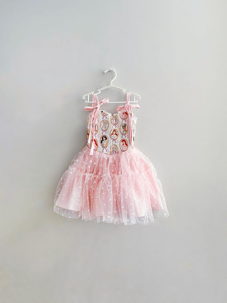 May include: A pink dress with a princess-themed bodice and a layered tulle skirt. The bodice features a repeating pattern of princess portraits. The dress has pink ribbon straps and is hanging on a white hanger.