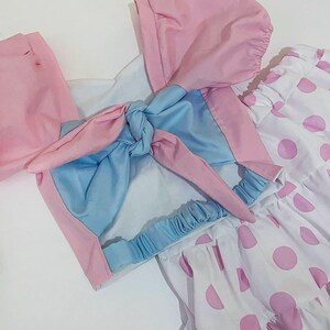 May include: A light pink and blue outfit with a matching bonnet and a white skirt with pink polka dots. The top has a bow detail and the skirt has a ruffled hem. The outfit is made of a lightweight fabric.