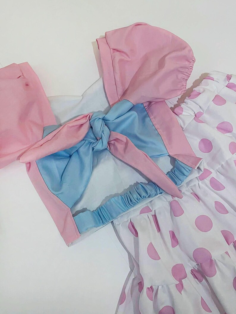 May include: A light pink and blue outfit with a matching bonnet and a white skirt with pink polka dots. The top has a bow detail and the skirt has a ruffled hem. The outfit is made of a lightweight fabric.
