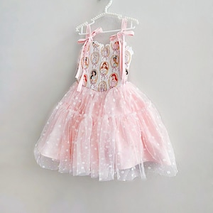 May include: A pink dress with a princess-themed bodice and a layered tulle skirt. The bodice features a repeating pattern of princess portraits in oval frames. The dress has pink ribbon straps and is hanging on a white hanger.