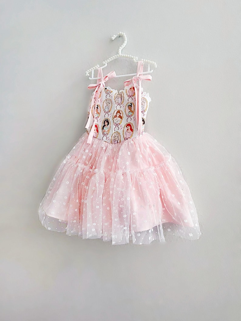 May include: A pink dress with a princess-themed bodice and a layered tulle skirt. The bodice features a repeating pattern of princess portraits in oval frames. The dress has pink ribbon straps and is hanging on a white hanger.