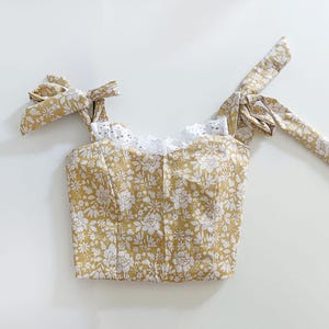 May include: A yellow floral crop top with white lace trim and tie straps. The top has a fitted bodice and a relaxed fit.
