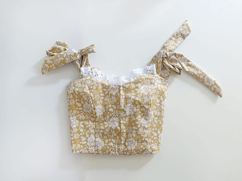 May include: A yellow floral crop top with white lace trim and tie straps. The top has a fitted bodice and a relaxed fit.
