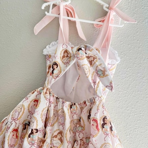 May include: A light pink dress with a pattern of princess portraits in oval frames. The dress has pink ribbon straps tied into bows and is hanging on a white hanger. The dress has a ruffled neckline and a bow in the back.
