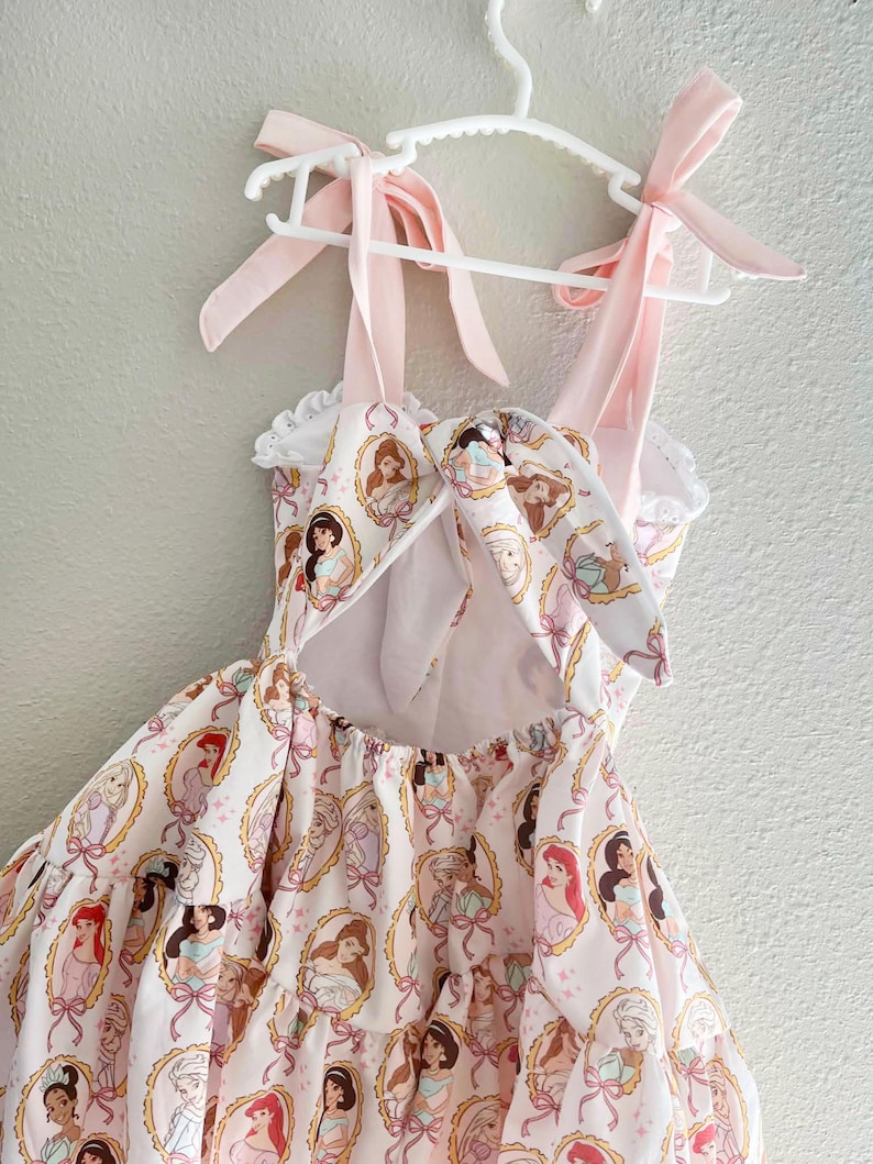 May include: A light pink dress with a pattern of princess portraits in oval frames. The dress has pink ribbon straps tied into bows and is hanging on a white hanger. The dress has a ruffled neckline and a bow in the back.