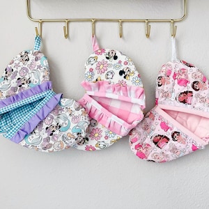 May include: Three hanging oven mitts with different patterns. One features a mermaid and Minnie Mouse print, another has a floral and Minnie Mouse design, and the last one showcases a princess theme. Each mitt has a ruffled edge and a loop for hanging.