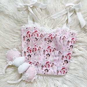 May include: A pink and white crop top with a print of Disney princesses. The top has white straps that tie at the shoulders and a white lace trim. The top is paired with pink and white mouse ears with a white bow.