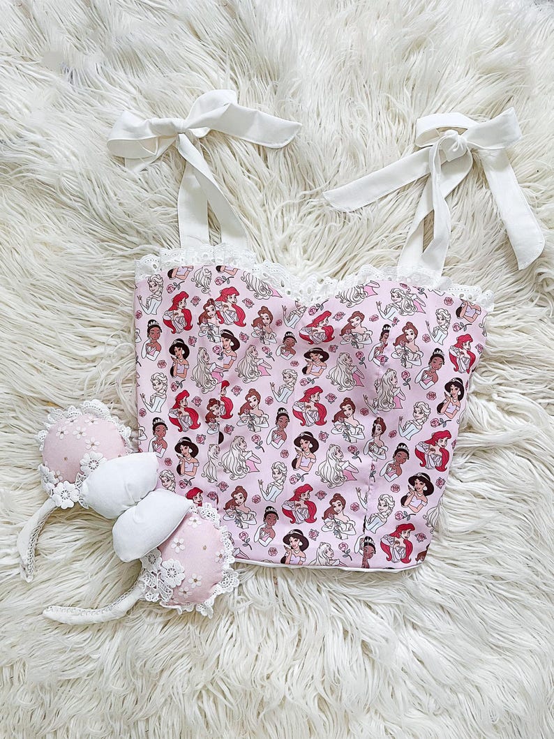 May include: A pink and white crop top with a print of Disney princesses. The top has white straps that tie at the shoulders and a white lace trim. The top is paired with pink and white mouse ears with a white bow.