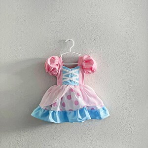 May include: A pink and blue dress with a white bodice and puff sleeves hangs on a white hanger. The dress features a pink skirt with large pink polka dots and a blue ruffled hem. The bodice has a crisscross design.