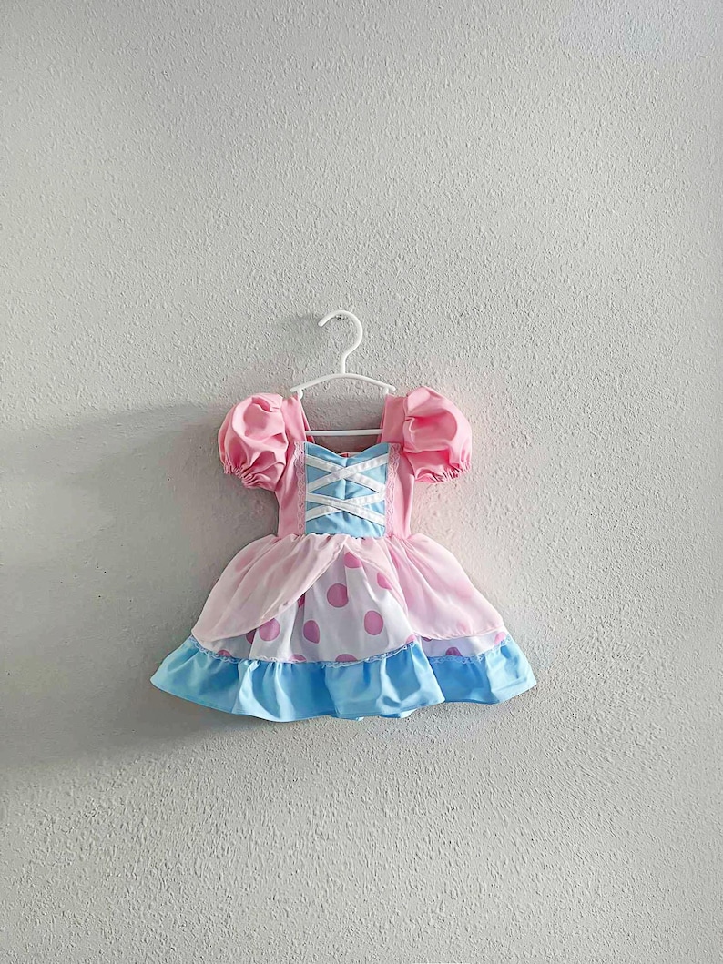 May include: A pink and blue dress with a white bodice and puff sleeves hangs on a white hanger. The dress features a pink skirt with large pink polka dots and a blue ruffled hem. The bodice has a crisscross design.