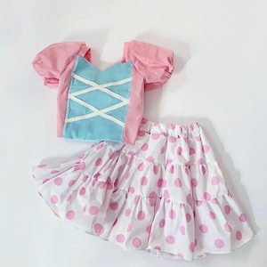 May include: A two-piece outfit featuring a pink and blue top and a white skirt with pink polka dots. The top has puffy sleeves and a corset-style front. The skirt is tiered.