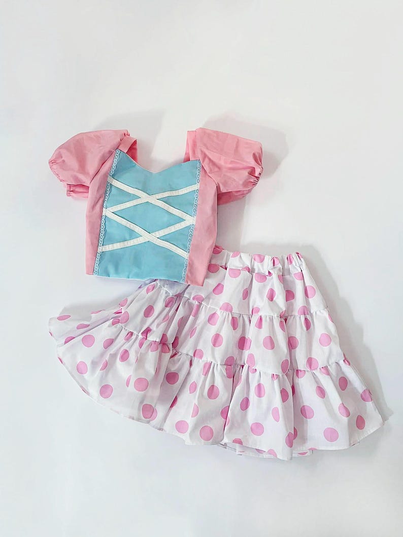 May include: A two-piece outfit featuring a pink and blue top and a white skirt with pink polka dots. The top has puffy sleeves and a corset-style front. The skirt is tiered.