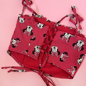 May include: Red crop top with a Minnie Mouse print and a lace-up back. The straps are tied at the shoulders.