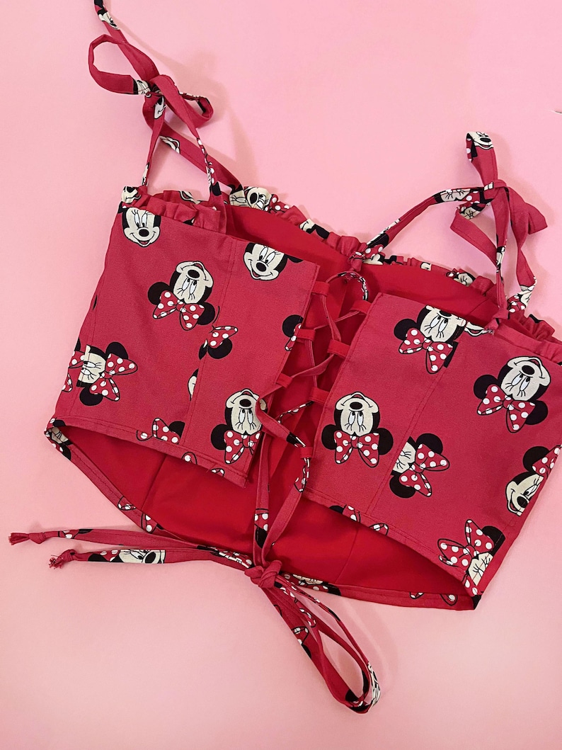 May include: Red crop top with a Minnie Mouse print and a lace-up back. The straps are tied at the shoulders.