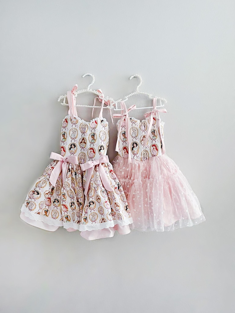May include: Two dresses hanging on white hangers. One dress is white with a pattern of princess portraits, pink bows, and a lace trim. The other is a pink tulle dress with white polka dots.
