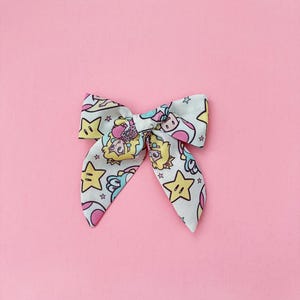 K&ouml;nnte beinhalten: Eine wei&szlig;e Schleife mit einem rosa, gelben und blauen Druck mit Comicfiguren und Sternen. Die Schleife ist gebunden und bereit zum Tragen.