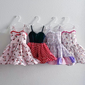 May include: Four sleeveless dresses hanging on white plastic hangers. The first is light pink with Minnie Mouse prints, the second has a black bodice and red polka-dot skirt, the third is lavender with character prints, and the fourth is white with colorful patterns.