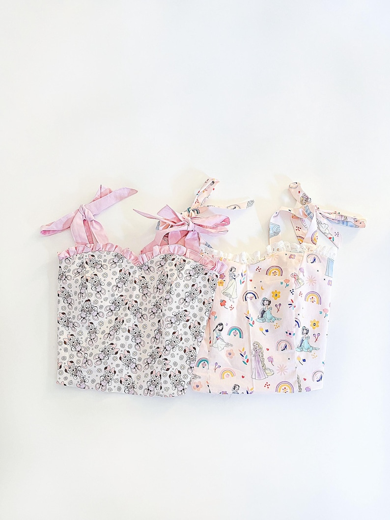 May include: Three pink and white sleeveless tops with different patterns. The first top has a small floral pattern with a brown and white design. The second top has a pink and white pattern with a rainbow and princesses. The third top has a pink and white pattern with a rainbow and princesses.