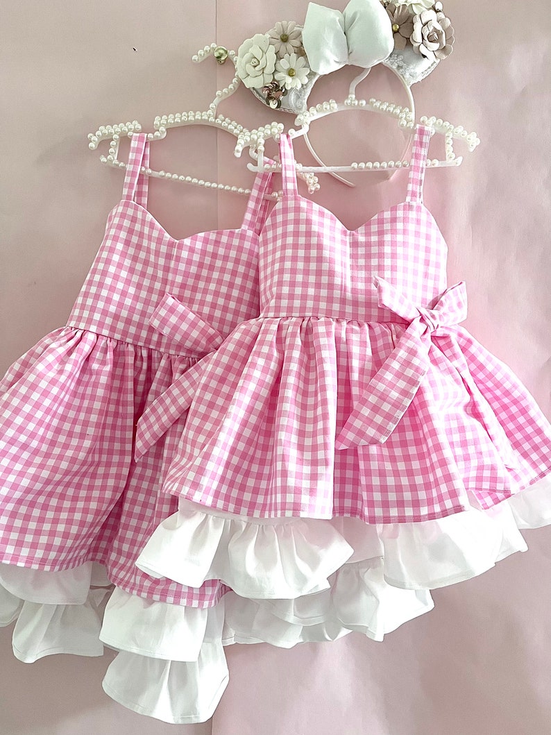 May include: Two pink and white gingham dresses with white ruffled layers. The dresses have a sweetheart neckline and are sleeveless. barbie dress
