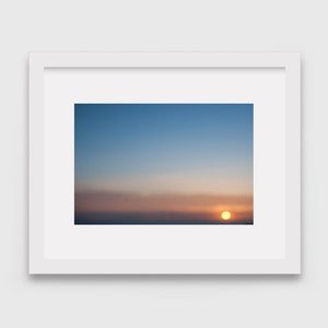 May include: A framed photograph of a sunset with a bright orange sun setting over a horizon of clouds. The sky is a gradient of blue, pink, and orange.