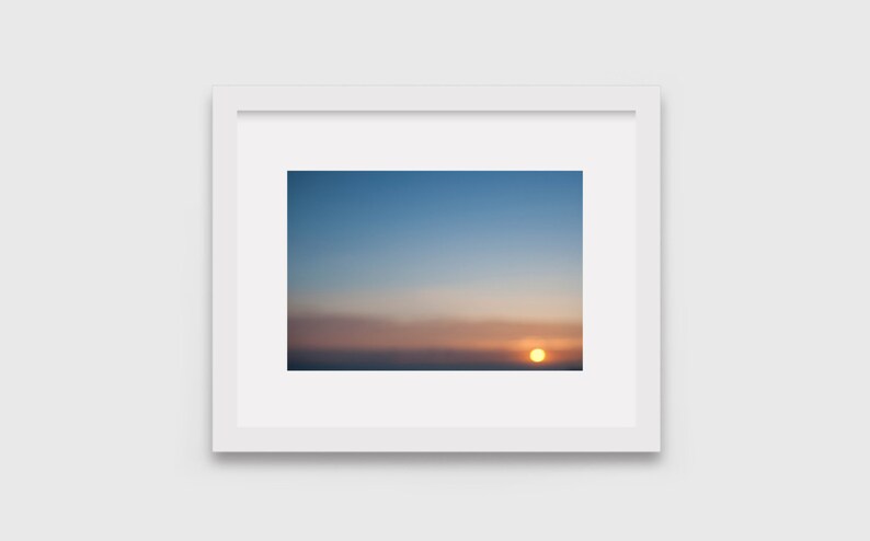 May include: A framed photograph of a sunset with a bright orange sun setting over a horizon of clouds. The sky is a gradient of blue, pink, and orange.