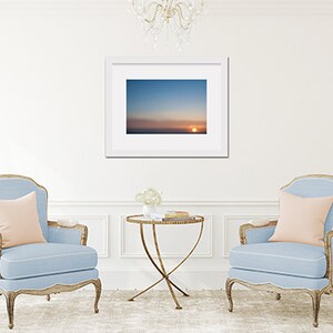 May include: Two light blue armchairs with pink pillows, a gold metal table with a glass top, and a framed photograph of a sunset over the ocean.