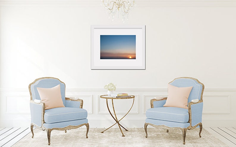 May include: Two light blue armchairs with pink pillows, a gold metal table with a glass top, and a framed photograph of a sunset over the ocean.